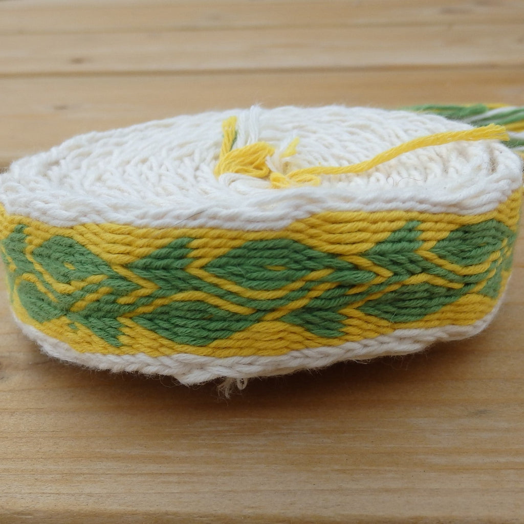 A handmade woven rope belt or waist wrap made from white, yellow, and green yarn, featuring a geometric leaf pattern, resting on a light wooden surface.