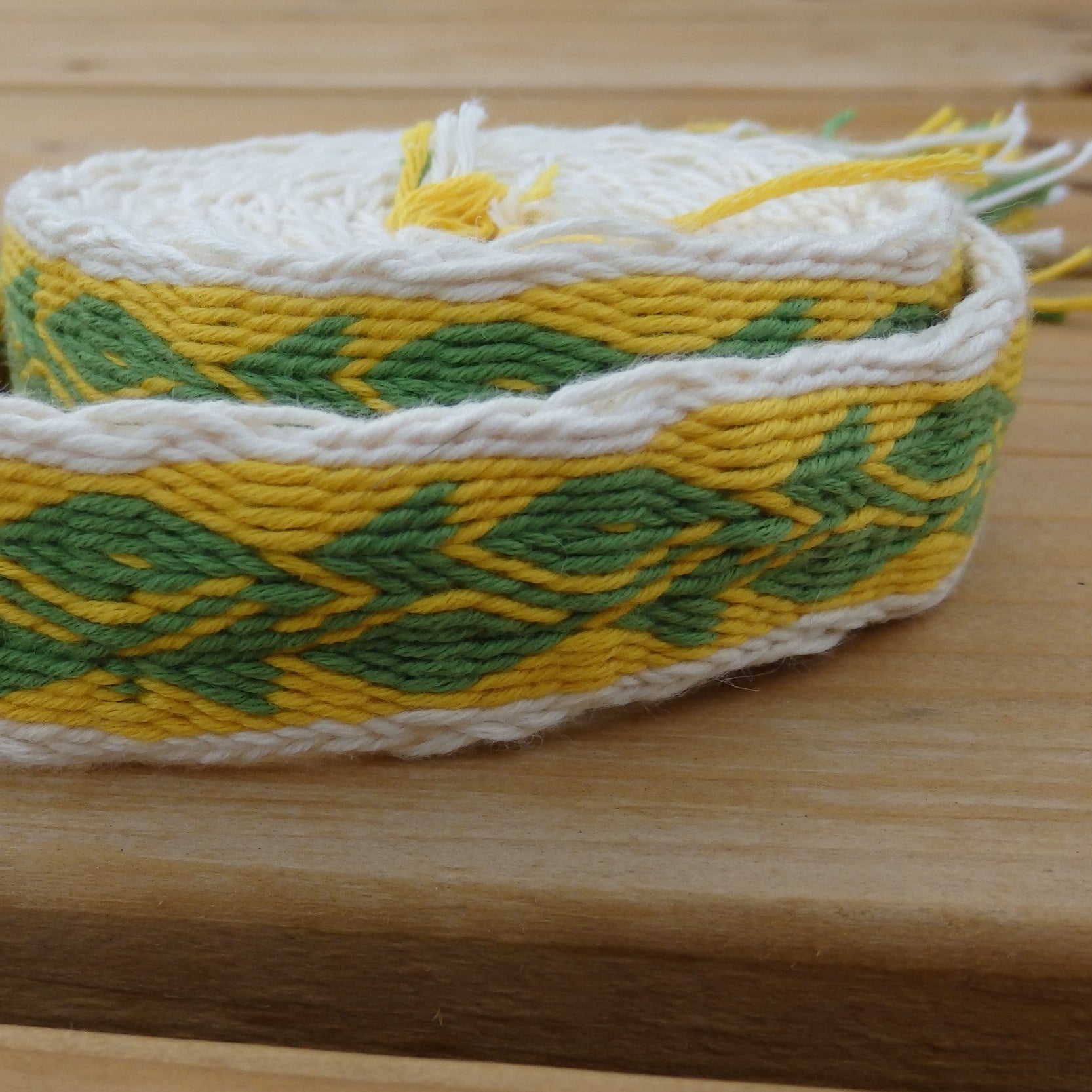 A handmade woven belt made of yellow, green, and white yarn with a geometric pattern, resting on a light wooden surface.