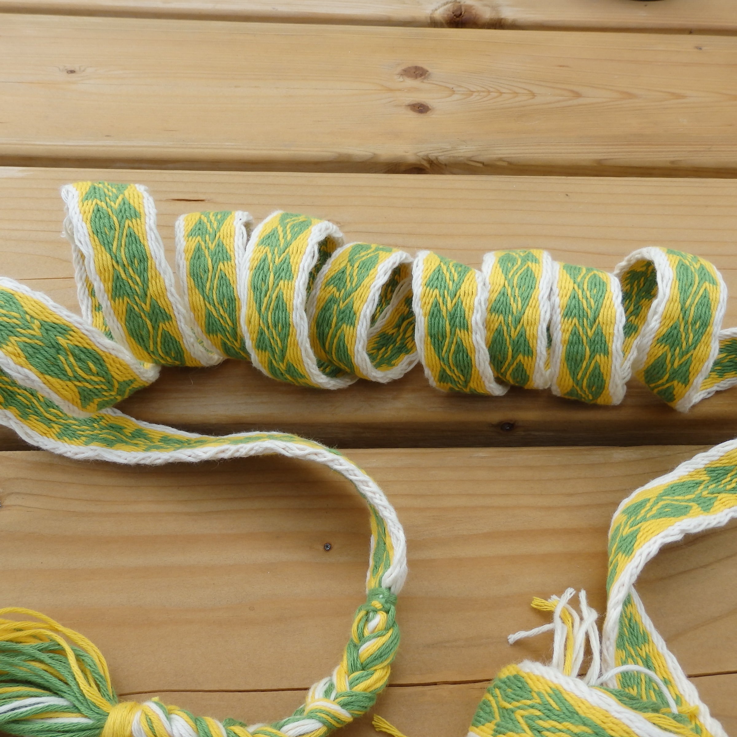 A braided wool scarf with a green and yellow leaf pattern on a white base, coiled on a light wood plank surface.