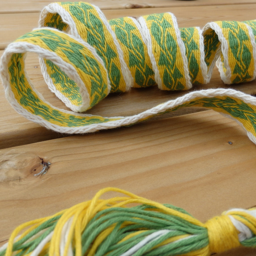A close-up of a braided rope or cord with a repeating green, yellow, and white pattern, laid on a light wood surface.