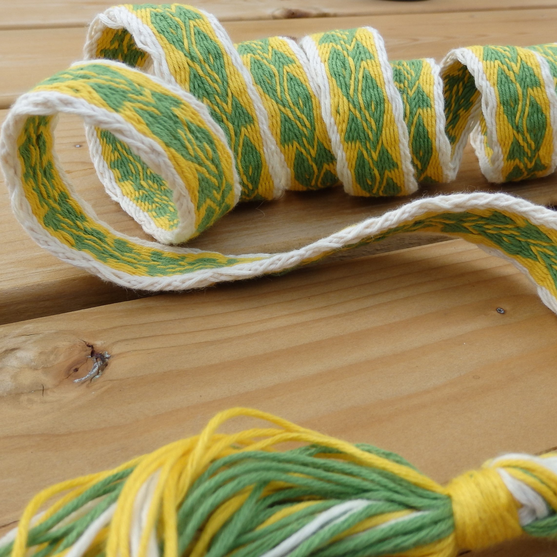 A close-up of a braided rope or cord with a repeating green, yellow, and white pattern, laid on a light wood surface.