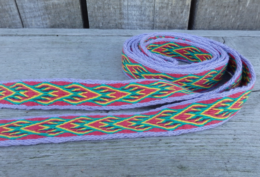 A woven decorative ribbon with a geometric pattern in red, green, yellow, and purple, coiled on a weathered gray wooden surface.
