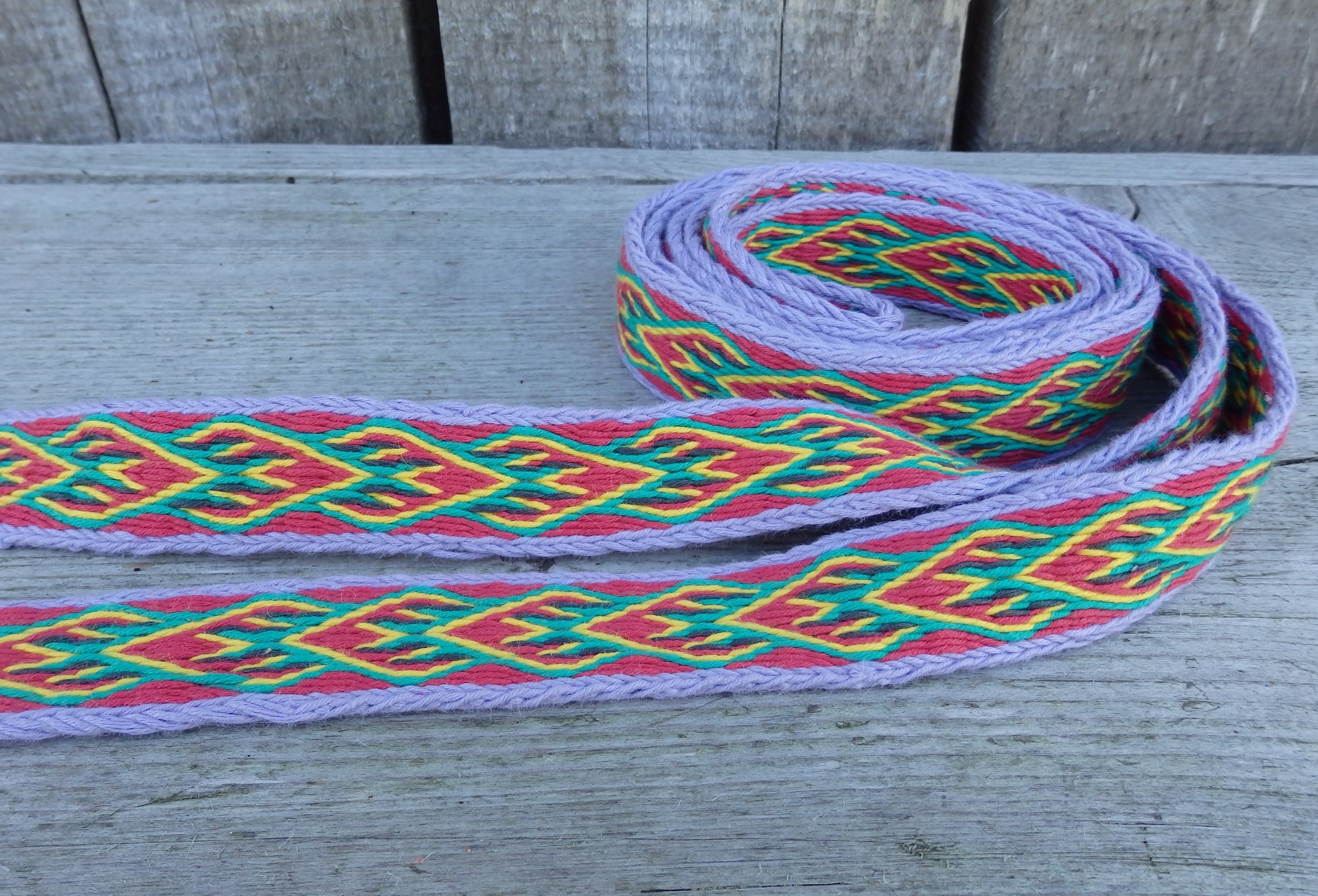 A woven decorative ribbon with a geometric pattern in red, green, yellow, and purple, coiled on a weathered gray wooden surface.