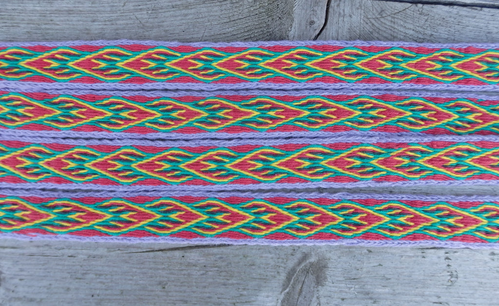 A close-up of a woven textile band with a colorful geometric pattern in red, green, yellow, and purple, laid flat on a weathered gray wooden surface.