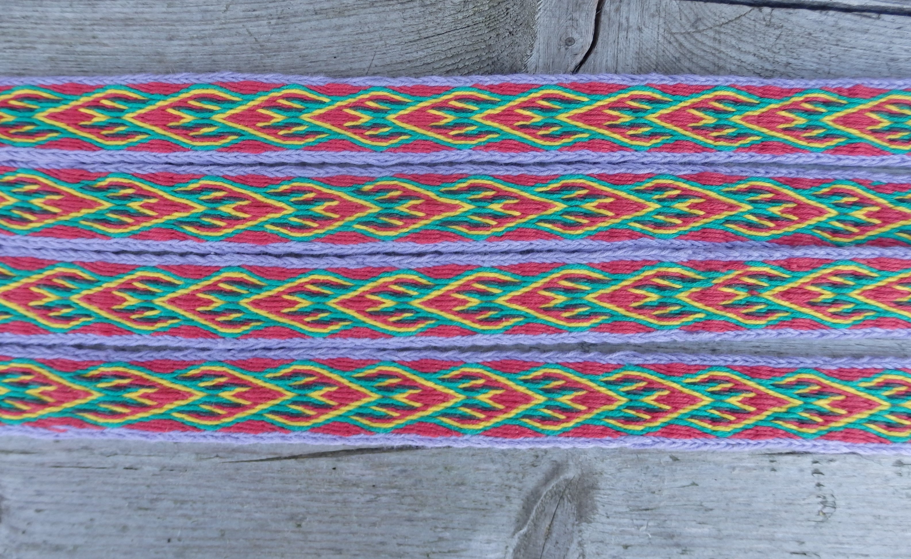 A close-up of a woven textile band with a colorful geometric pattern in red, green, yellow, and purple, laid flat on a weathered gray wooden surface.