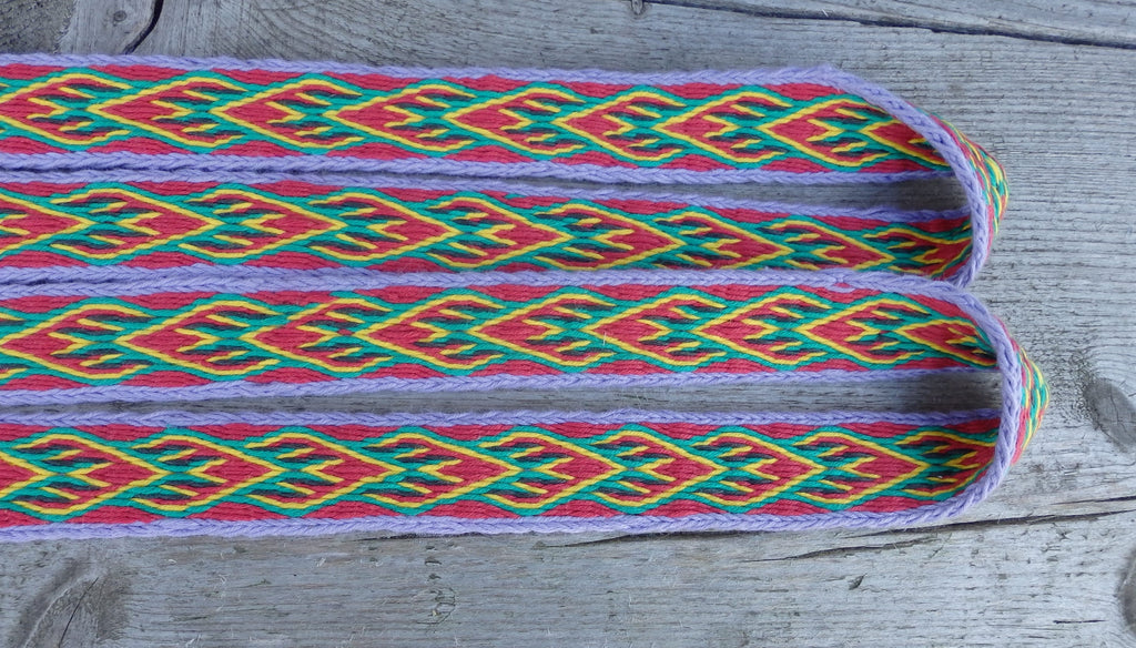 A close-up of a colorful woven textile or strap with a geometric pattern in red, green, yellow, and purple, laid flat on a weathered gray wooden surface.