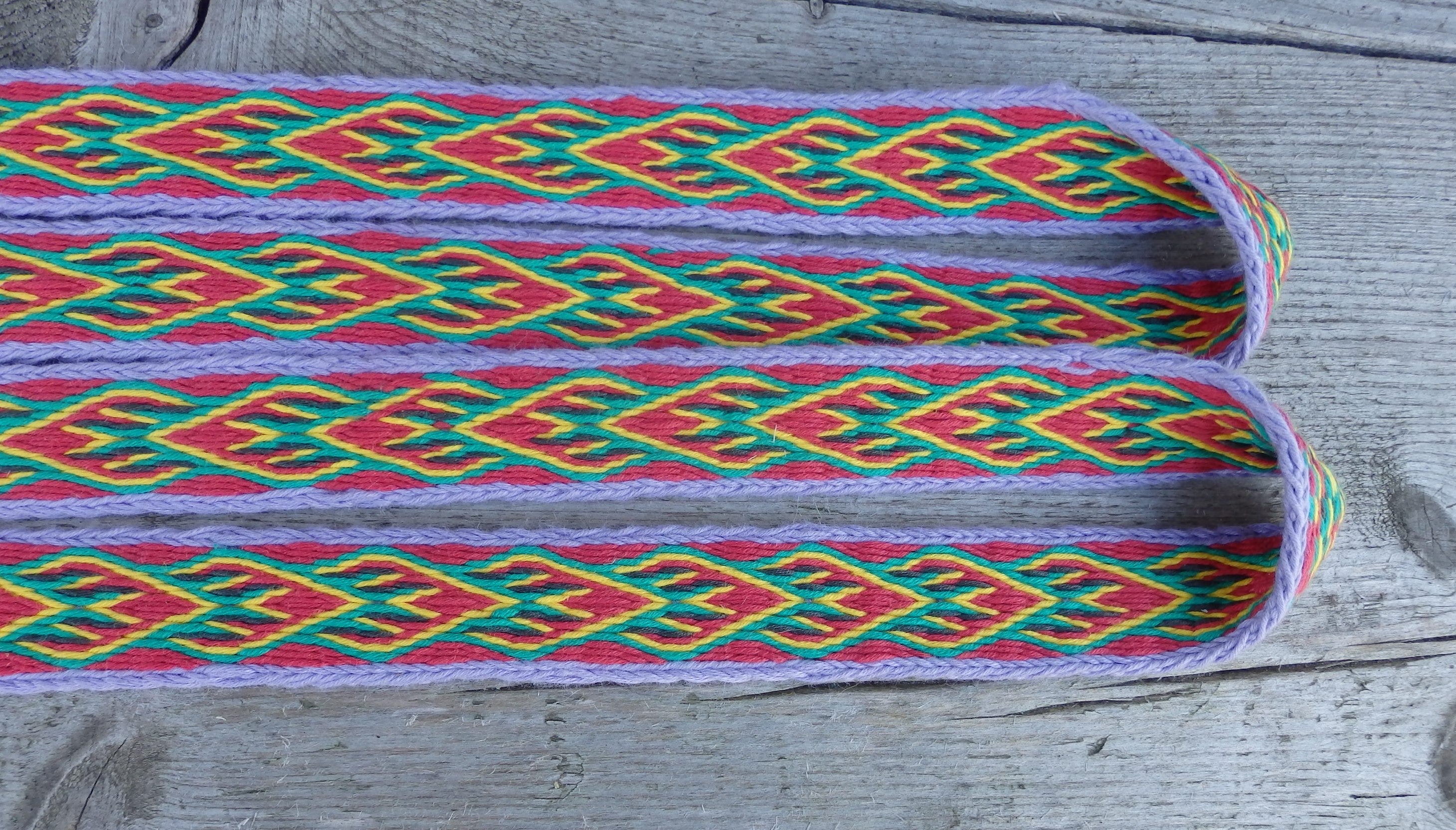 A close-up of a colorful woven textile or strap with a geometric pattern in red, green, yellow, and purple, laid flat on a weathered gray wooden surface.