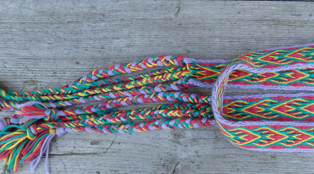A close-up of a colorful braided textile accessory, likely a braided strap or decorative trim, laid on a weathered gray wooden surface. The braid and woven pattern contain multiple bright colors including pink, green, yellow, orange, and purple.