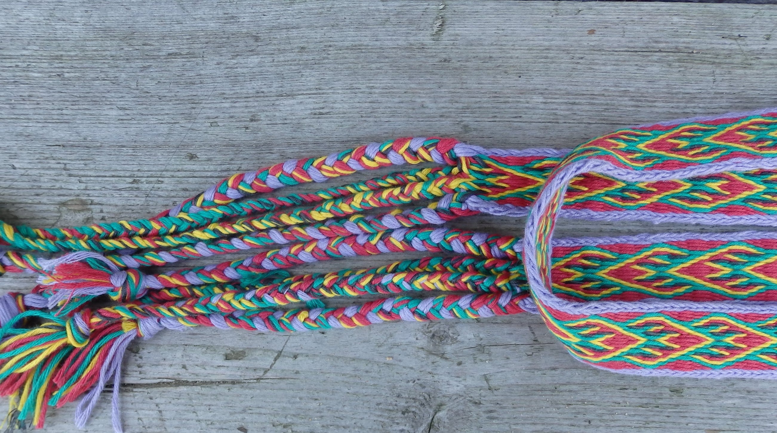 A close-up of a colorful braided textile accessory, likely a braided strap or decorative trim, laid on a weathered gray wooden surface. The braid and woven pattern contain multiple bright colors including pink, green, yellow, orange, and purple.