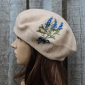 Beige beret with embroidered blue and green design on a mannequin head against a striped background