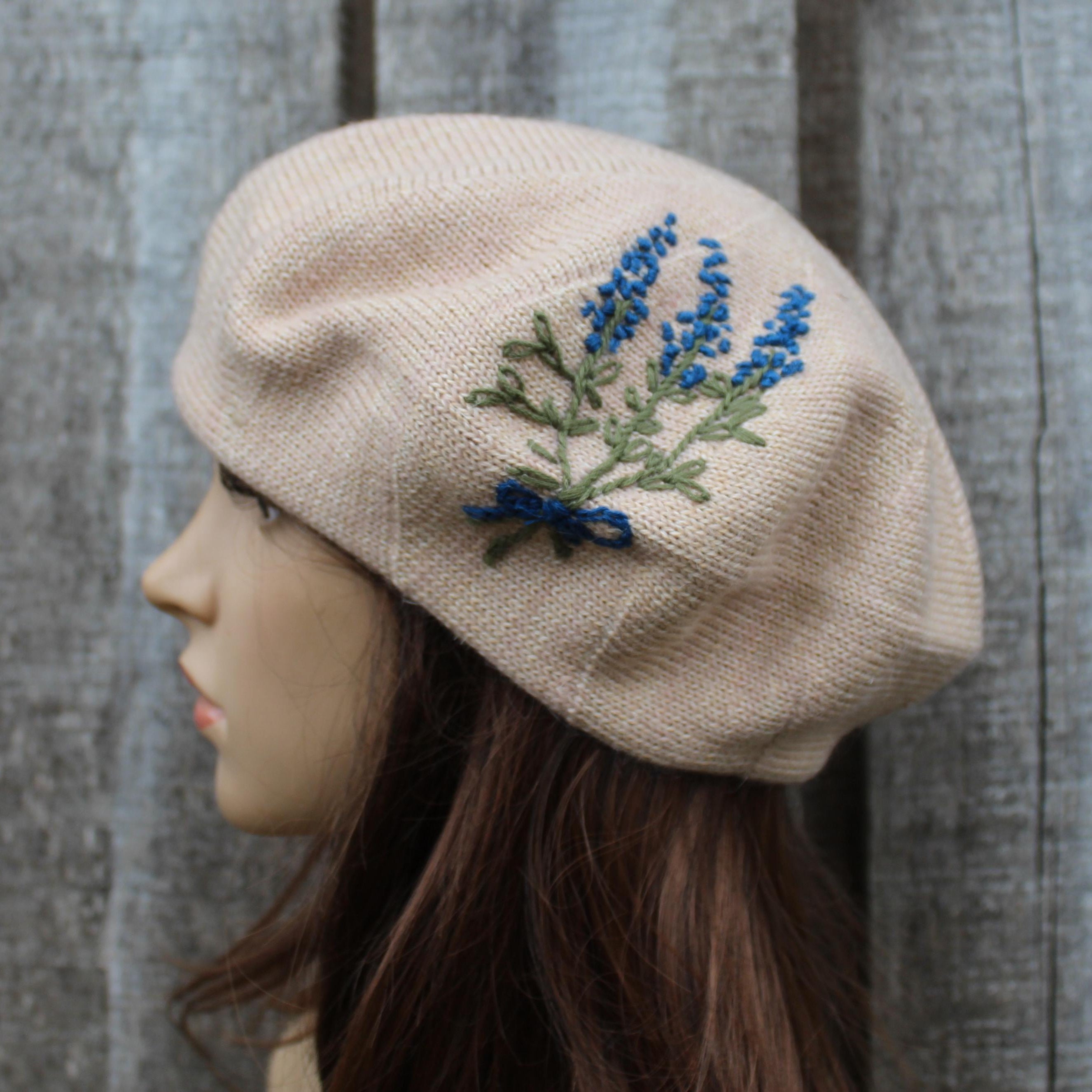 Beige beret with embroidered blue and green design on a mannequin head against a striped background