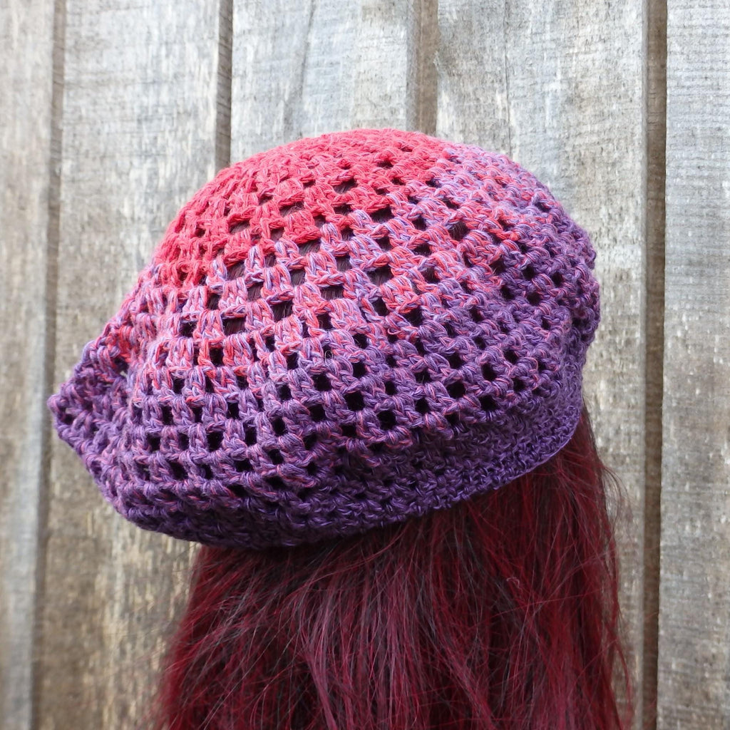 Purple and pink crocheted hat worn by a person with red hair against a wooden background